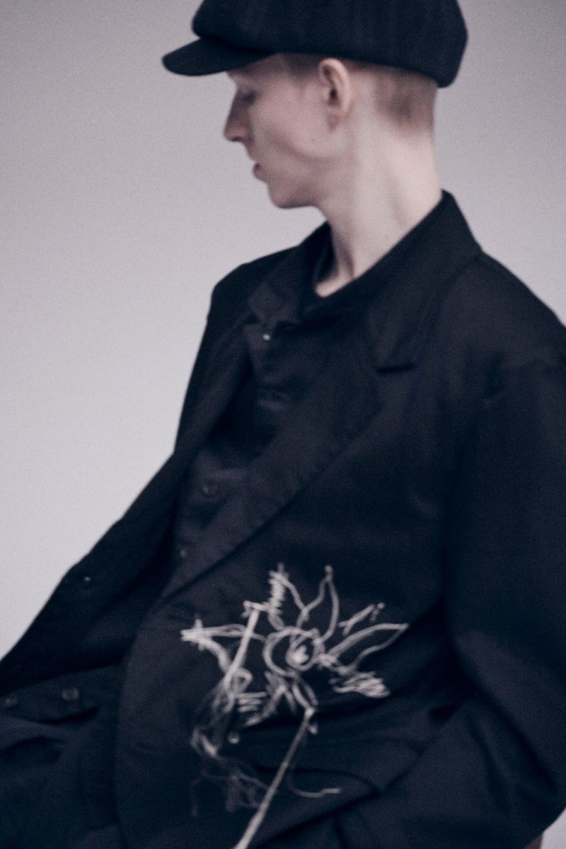 Headshot of a fair-skinned person in a black blazer with a floral design, wearing a cap, posing against a neutral backdrop.
