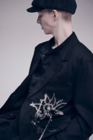 Headshot of a fair-skinned person in a black blazer with a floral design, wearing a cap, posing against a neutral backdrop.