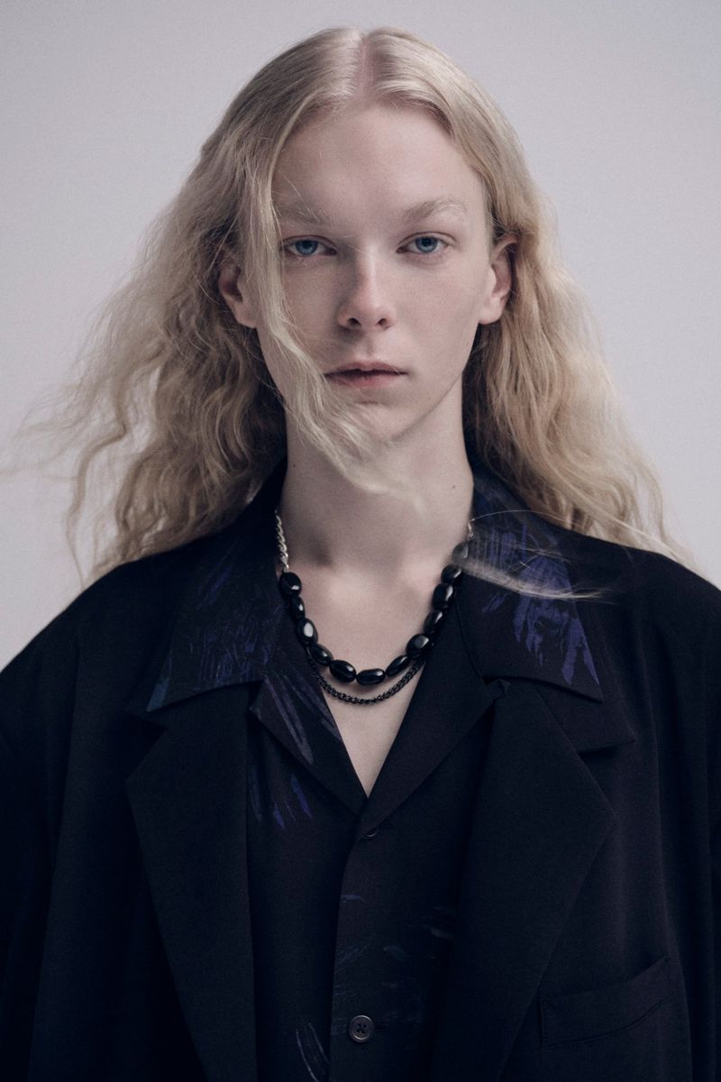 Close-up portrait of an adult female with long blonde hair, wearing a dark shirt and jacket, accessorized with a beaded necklace, set against a grey backdrop.