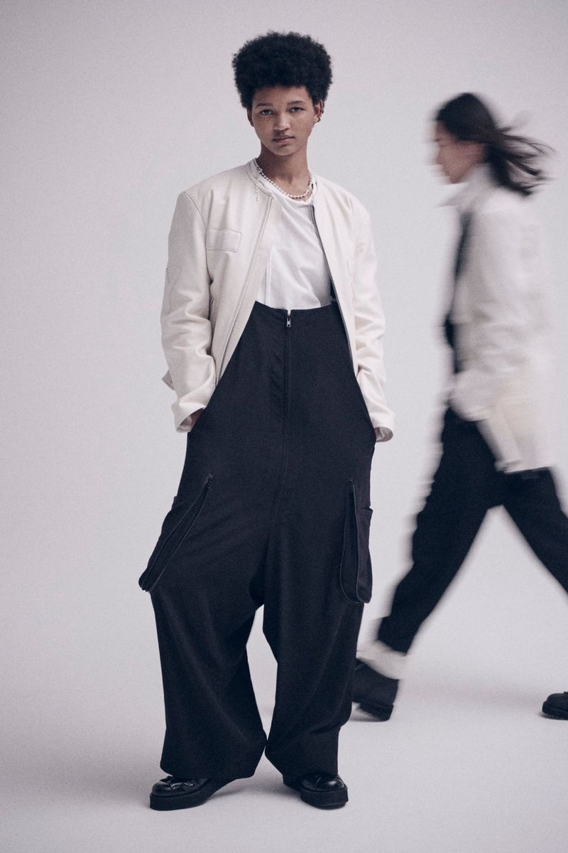 Two fashion models pose in a photo studio. One model is in focus, wearing a white jacket and black pants with a neutral expression.