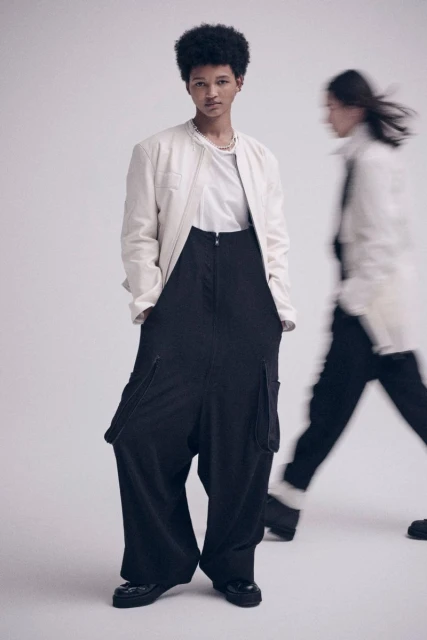 Two fashion models pose in a photo studio. One model is in focus, wearing a white jacket and black pants with a neutral expression.