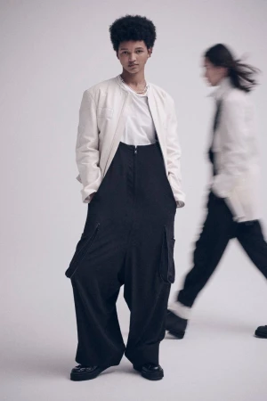 Two fashion models pose in a photo studio. One model is in focus, wearing a white jacket and black pants with a neutral expression.