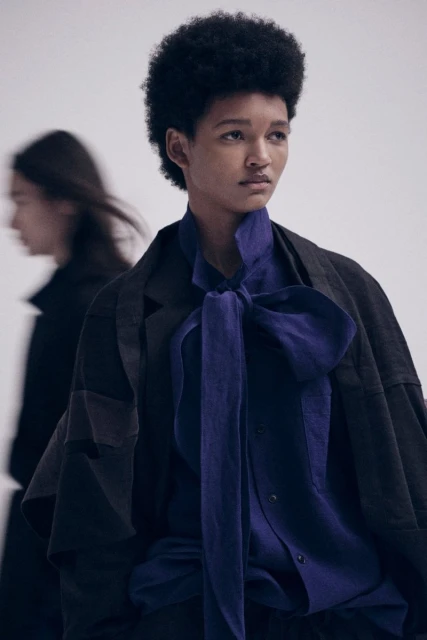 Fashion editorial shot of a Black woman in an oversized coat with a large bow tie standing in a bright studio, with another model blurred.