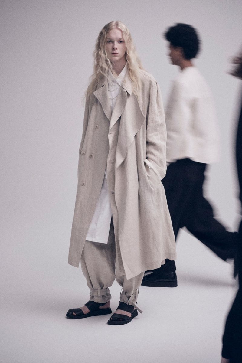 Fashion shot of a fair-skinned adult model in a trench coat, baggy pants, and sandals, standing in front of blurred people in a photo studio.
