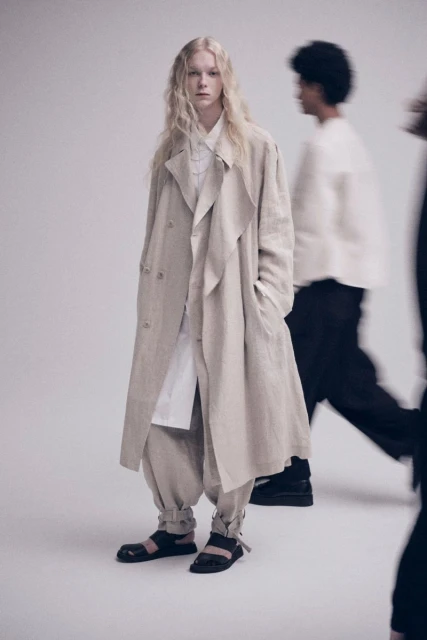 Fashion shot of a fair-skinned adult model in a trench coat, baggy pants, and sandals, standing in front of blurred people in a photo studio.
