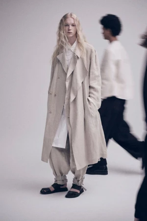 Model with long blonde hair wearing a beige coat and strapped sandals against a seamless white studio background.