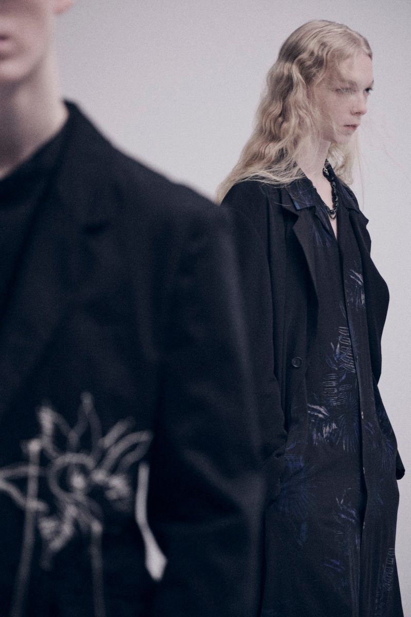 Two fashion models in black textured overcoats and black shirts in a studio. One model is fair-haired, one is partially out of view.