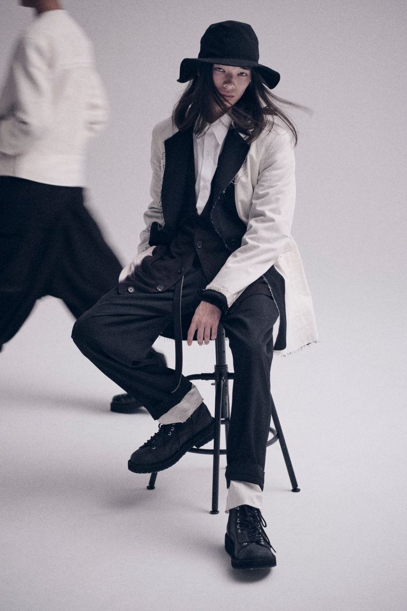 A person wearing a white jacket and black pants sitting on a stool in a studio, wearing a black hat. Another person stands blurred in the background.