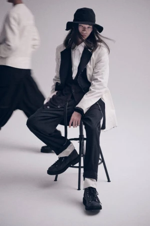 A person wearing a white jacket and black pants sitting on a stool in a studio, wearing a black hat. Another person stands blurred in the background.