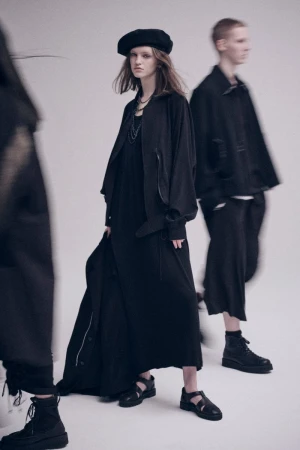 Three people in black clothing walking in a studio: an adult woman in a dress and jacket is in focus; two more are blurred.