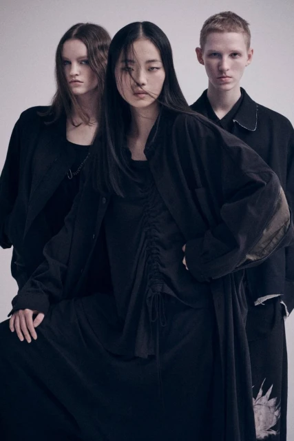 Three fashion models in dark clothing pose indoors against a neutral background, showcasing contemporary and minimalist style in a studio setting.