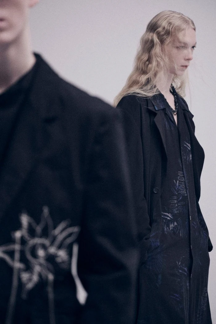 Model with long wavy blonde hair wearing a black coat standing behind another model in an embroidered dark jacket.