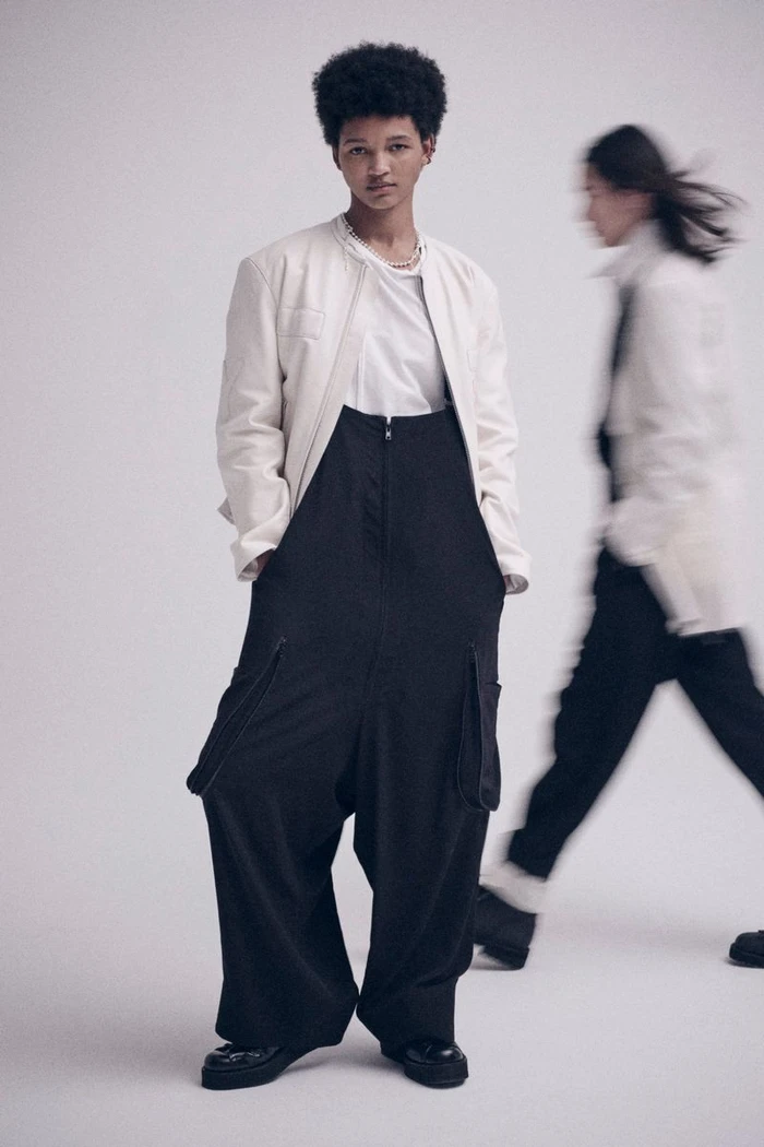 Model wearing a white jacket and black cargo overalls standing against a plain white studio background.