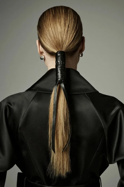 Woman with blonde hair styled in a low ponytail wrapped with black ribbon, wearing a black satin coat against a grey background.