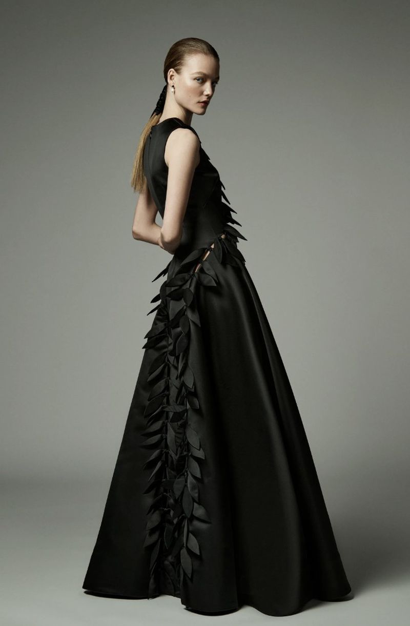 A fair-skinned woman poses in a full-length black gown with leaf accents against a neutral backdrop in a fashion forward studio portrait.
