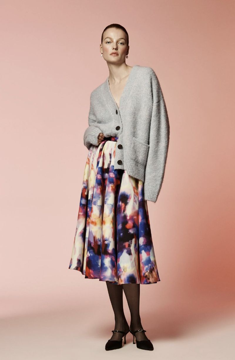 Full shot of a woman posing in a grey cardigan and a colorful patterned skirt against a light pink background in a fashion editorial.
