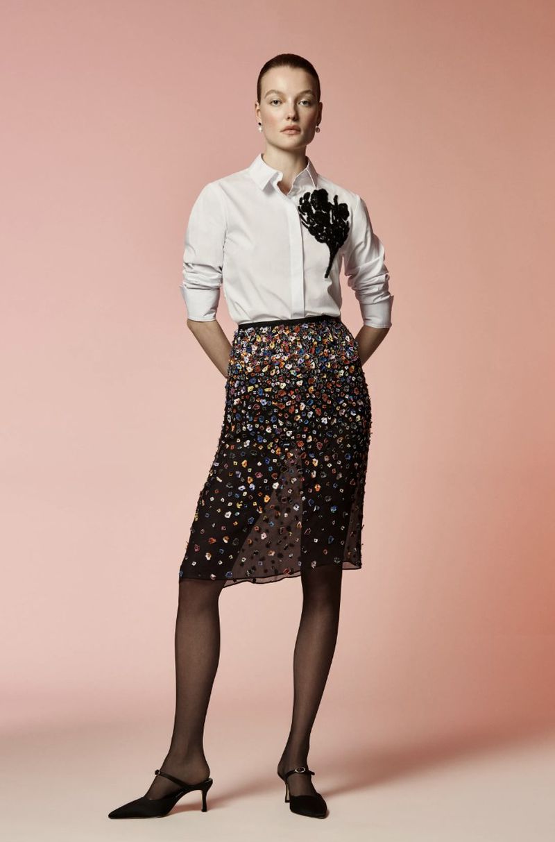 A woman stands in a studio wearing a white button-down shirt, semi-sheer skirt, black tights, and heels against a gradient pink backdrop.