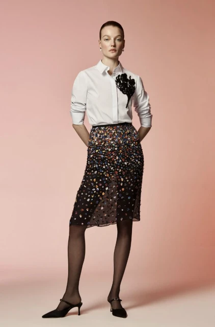 A woman stands in a studio wearing a white button-down shirt, semi-sheer skirt, black tights, and heels against a gradient pink backdrop.