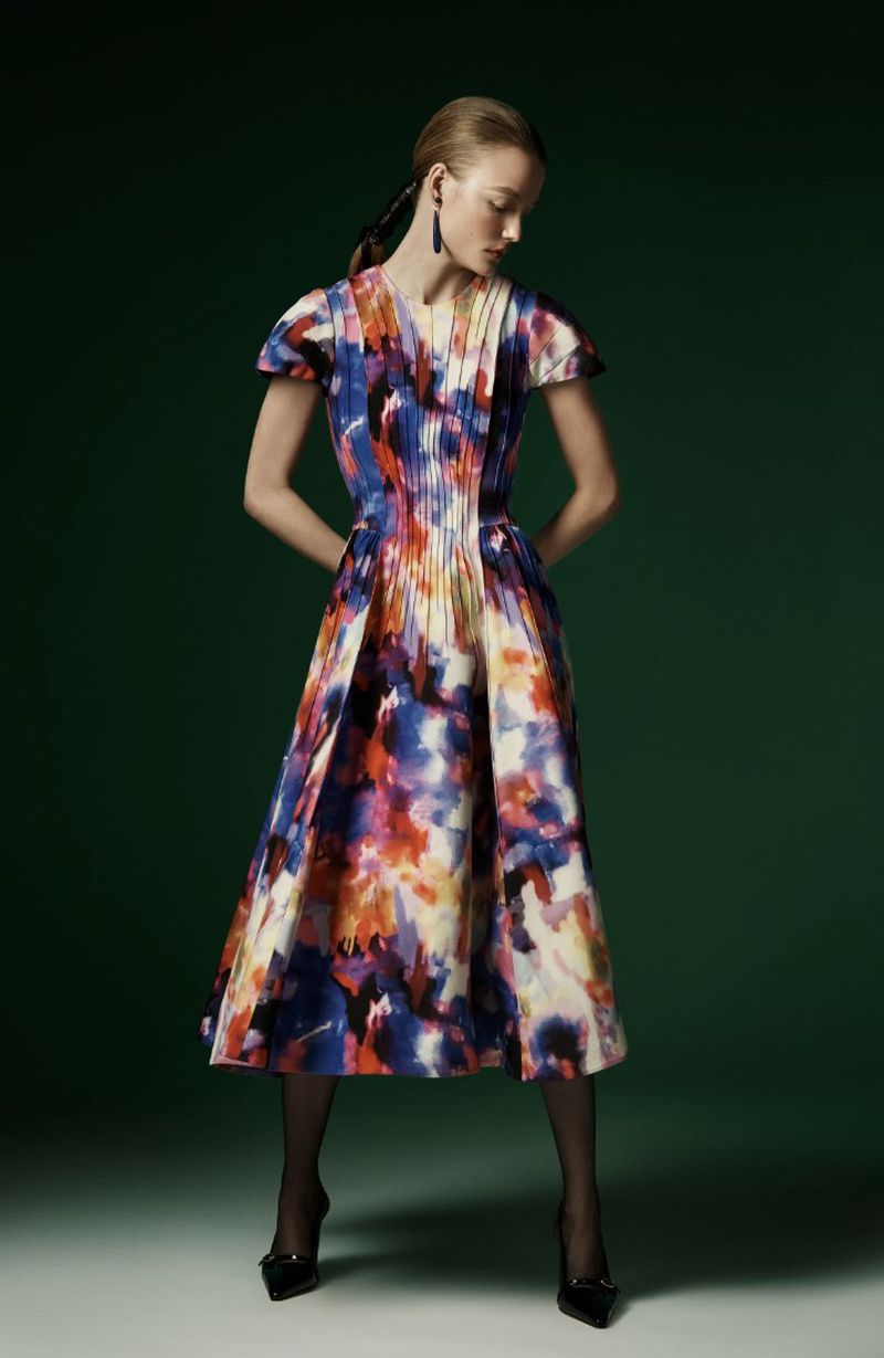 Full-length studio shot of an adult woman in a vibrant, patterned midi dress, black tights, and heeled shoes, set against a green backdrop.