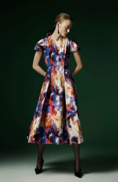 Full-length studio shot of an adult woman in a vibrant, patterned midi dress, black tights, and heeled shoes, set against a green backdrop.