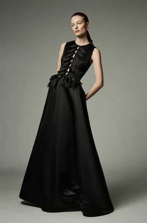 Model Roos Abels posing in a sleeveless black floor-length gown with leaf appliqué details against a neutral grey studio backdrop.