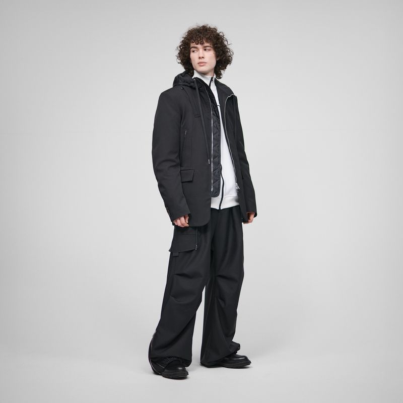 Model wearing a layered black jacket and wide-leg black cargo trousers against a neutral grey studio backdrop.