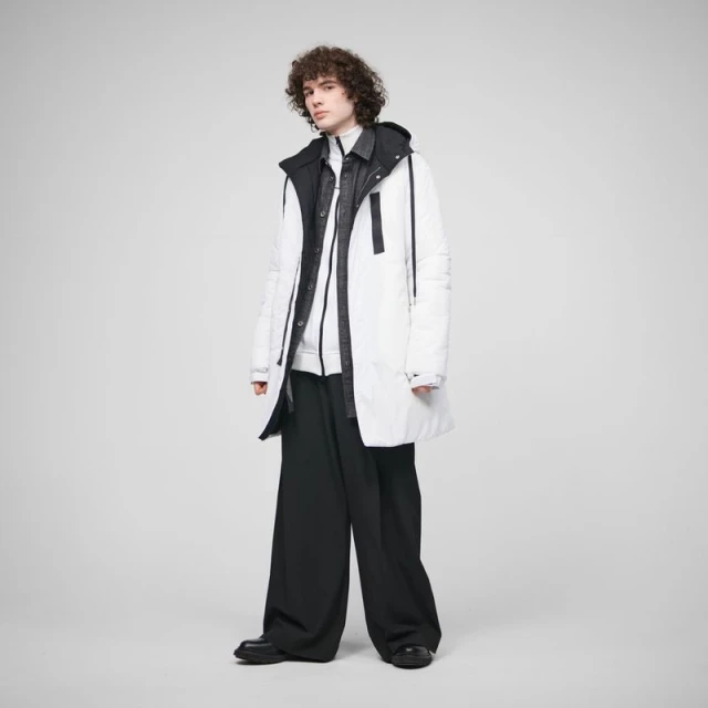 A young adult male model with curly brown hair poses in a white puffer coat and black pants against a light grey studio backdrop.