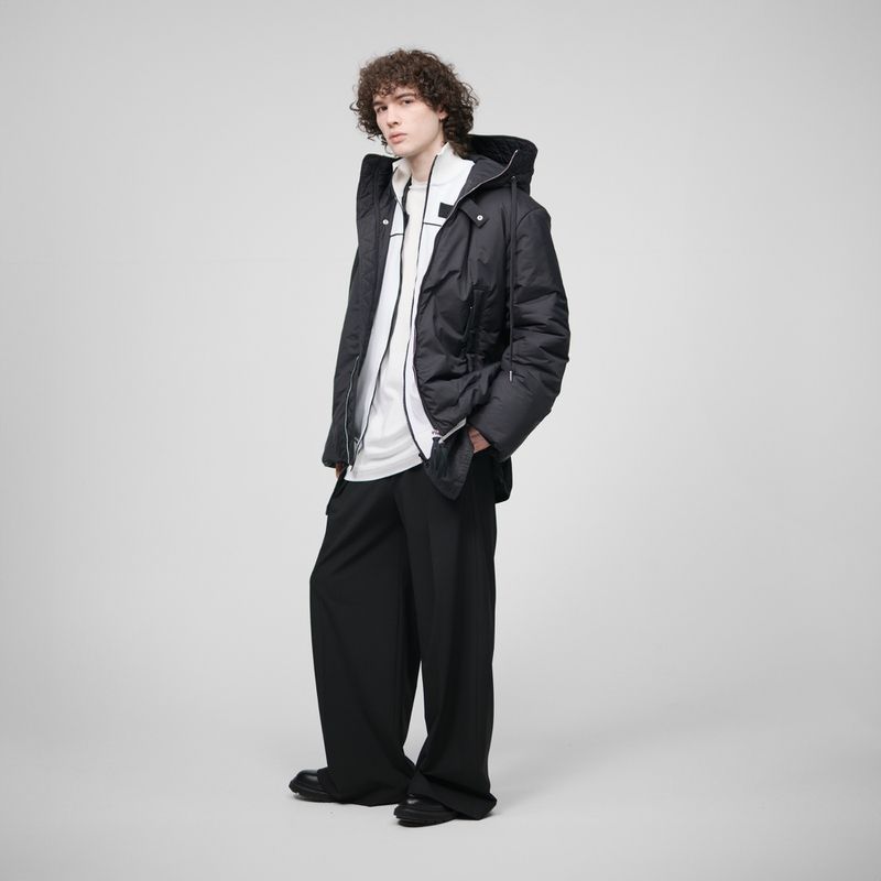 Young man with curly hair in a puffy black jacket, white top, and baggy black pants stands in a full shot against a neutral background.