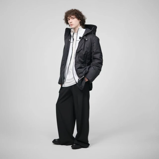 Young man with curly hair in a puffy black jacket, white top, and baggy black pants stands in a full shot against a neutral background.