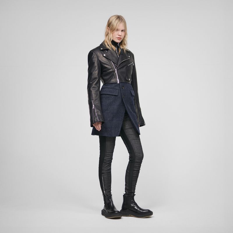Full-length shot of a blonde adult fashion model wearing a black leather jacket, asymmetrical skirt, dark patterned leggings, and boots.