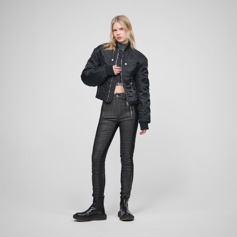 Woman posing in a black bomber jacket and dark denim jeans against a white background in a full studio shot.