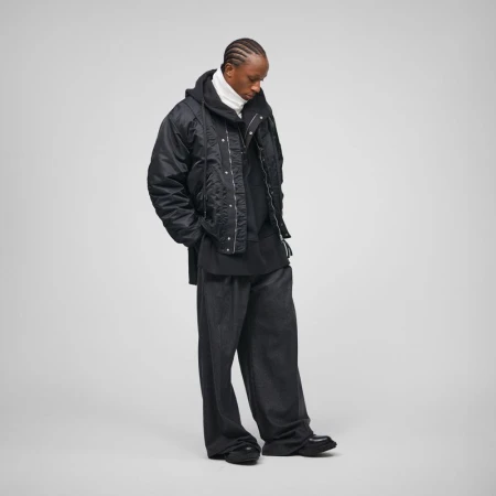Full-length fashion shot of a Black male model wearing an unzipped black bomber jacket, hooded sweatshirt, oversized denim pants, and black shoes.