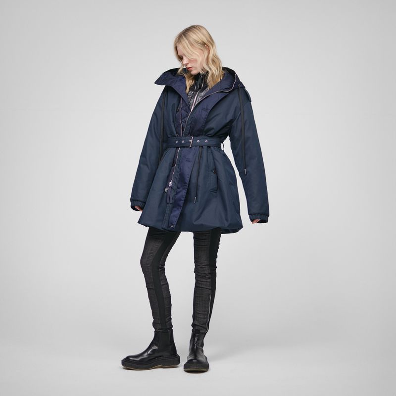 Fashion shot of a fair-skinned woman wearing a belted navy blue coat with black leggings and boots against a grey backdrop.  