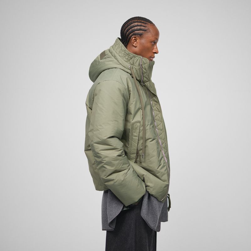 Profile shot of an adult male in a hooded olive green puffer jacket and gray sweater against a neutral background, showcasing contemporary fashion.