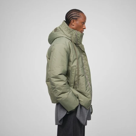 Profile shot of an adult male in a hooded olive green puffer jacket and gray sweater against a neutral background, showcasing contemporary fashion.