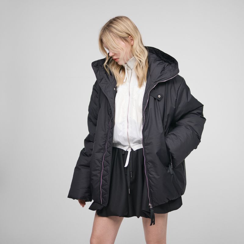 A blond-haired model is wearing a black hooded puffer jacket and short black skirt against a plain gray studio background.
