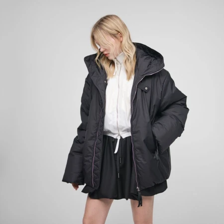 A blond-haired model is wearing a black hooded puffer jacket and short black skirt against a plain gray studio background. 