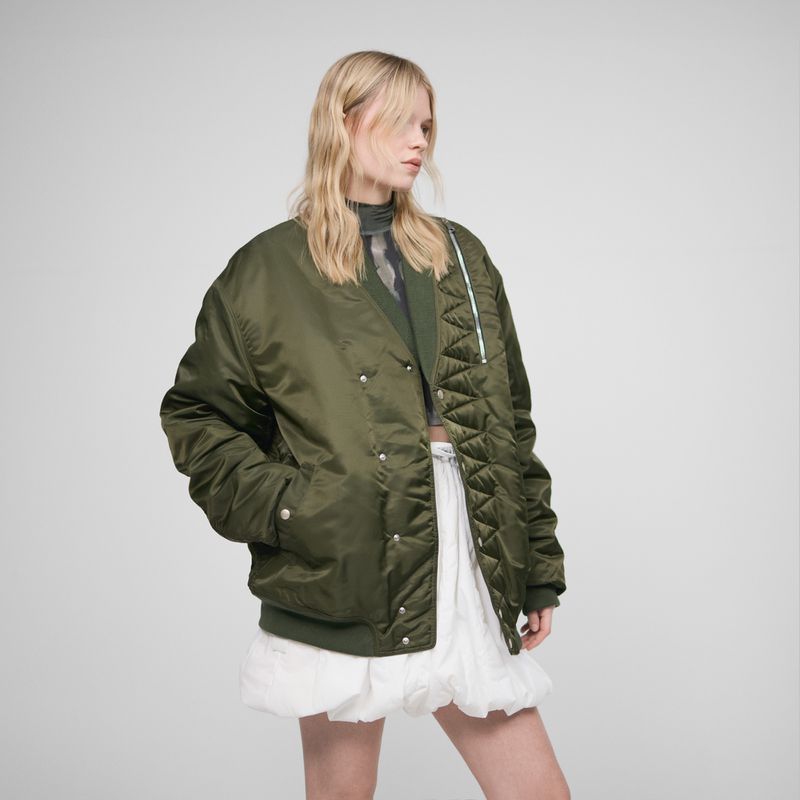 Fashion photo of a blonde model in an olive green bomber jacket with a white mini skirt, posing against a neutral grey backdrop.