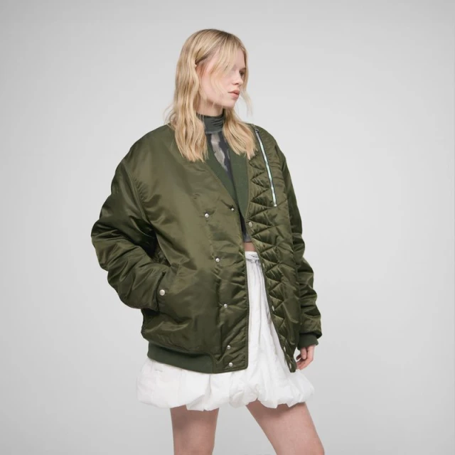 Fashion photo of a blonde model in an olive green bomber jacket with a white mini skirt, posing against a neutral grey backdrop.