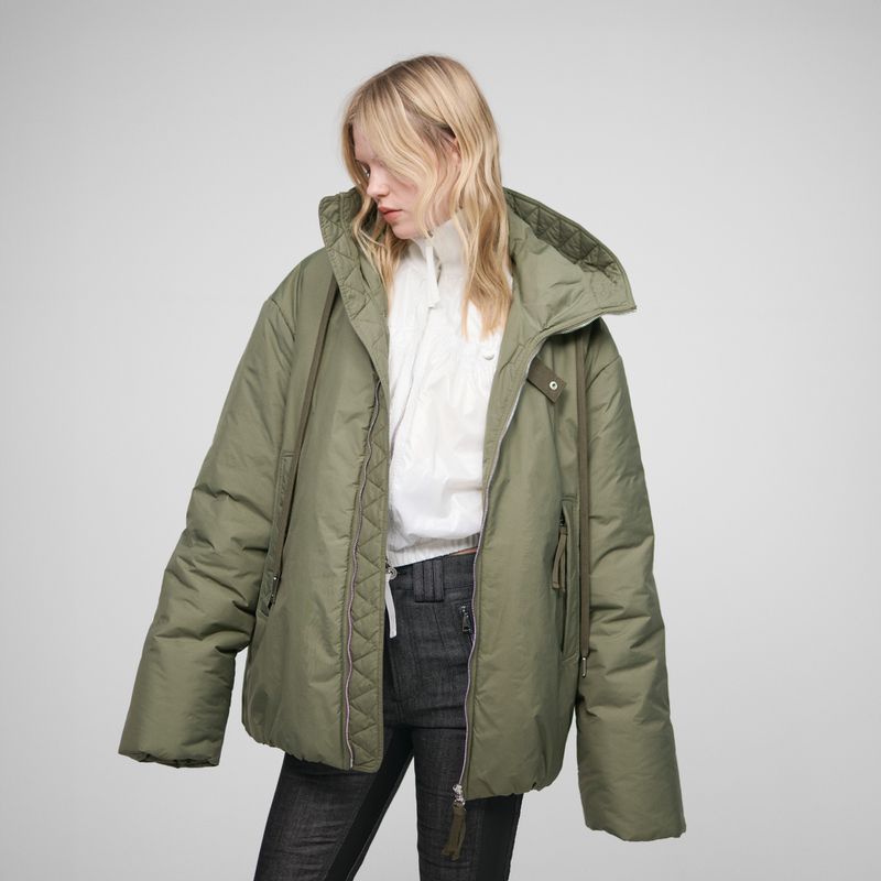 A woman with blonde hair poses in a studio with an olive green puffer jacket, a white top, and dark jeans against a neutral background.