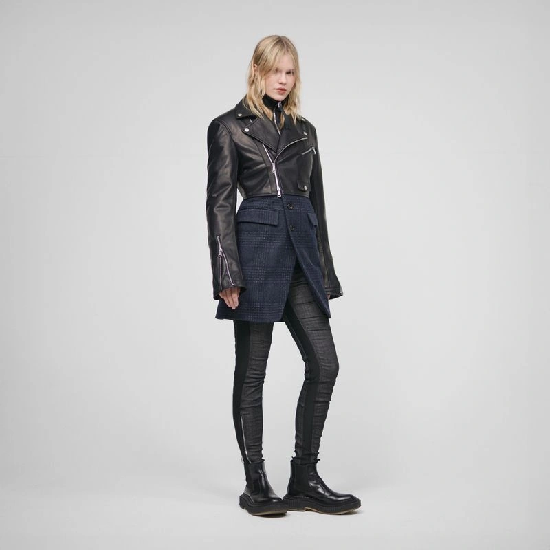 Model wearing a black leather cropped jacket and patterned skirt over dark trousers against a neutral grey studio backdrop.