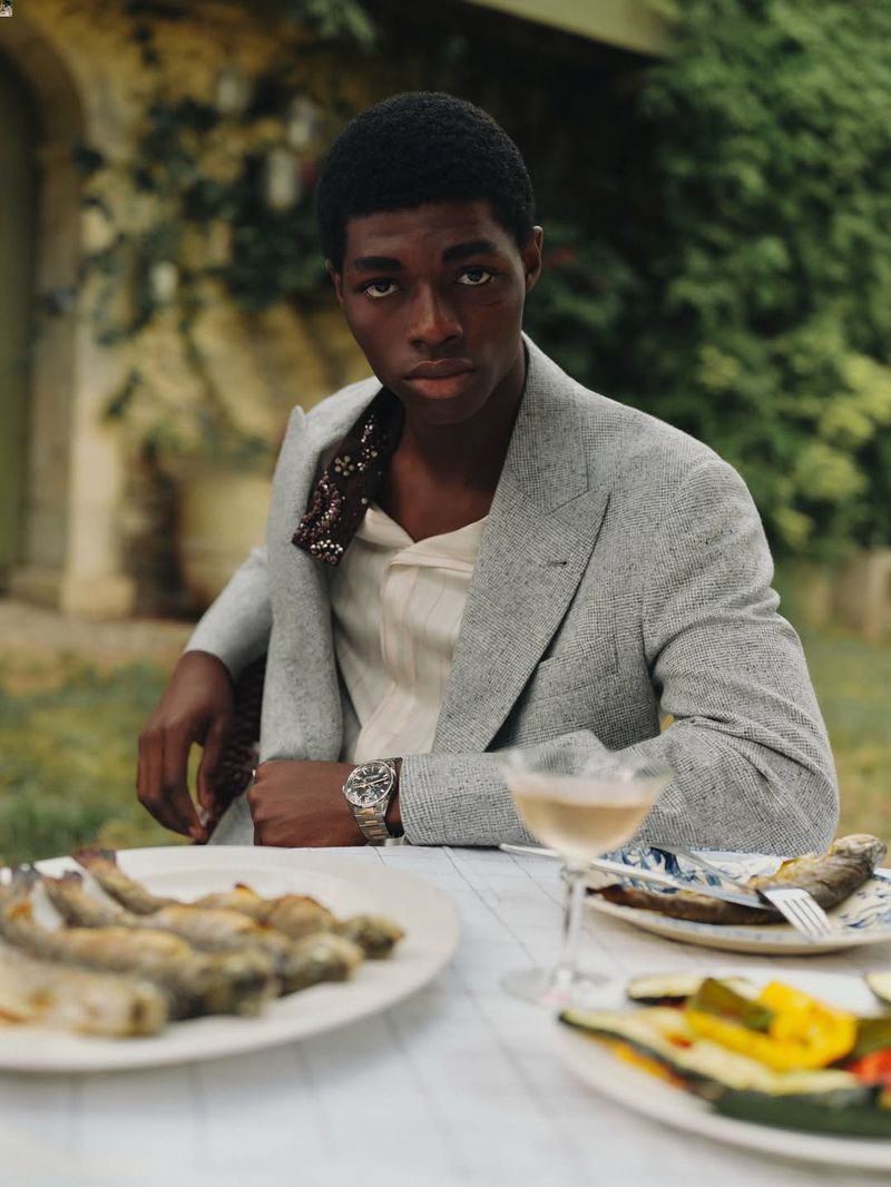 Portrait of a Black male model in a grey suit jacket seated at a table with fish, wine, and green foliage in the background.