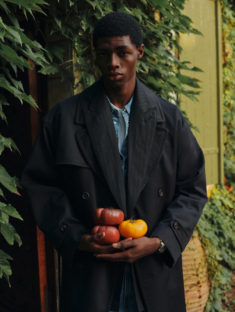Dark-skinned male model in overcoat holds colorful tomatoes outdoors amid greenery, creating a natural, fashion-forward visual.