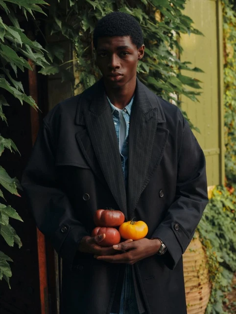 Dark-skinned male model in overcoat holds colorful tomatoes outdoors amid greenery, creating a natural, fashion-forward visual.