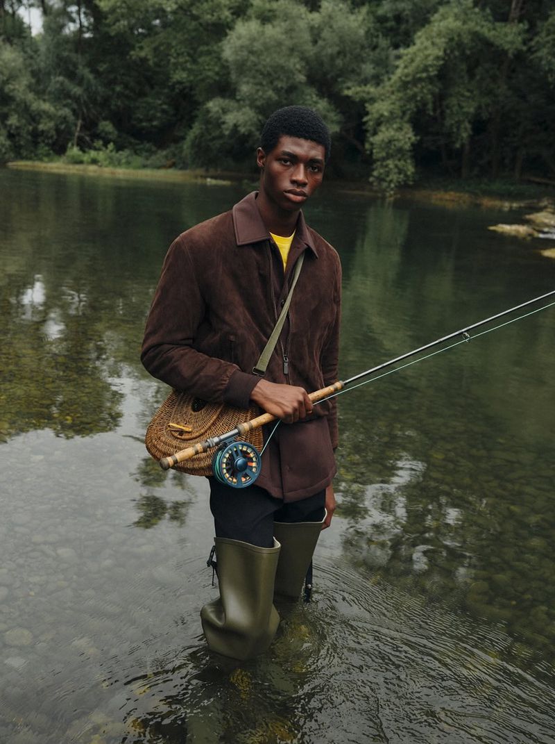 Man in a brown jacket and waders stands in a river holding a fishing rod with trees in the background, photographed at eye-level.