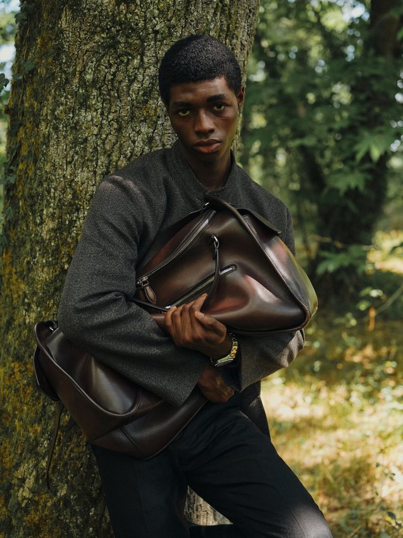 Man in grey jacket holds brown leather bag in forest. Portrait of adult male with brown hair and dark clothing in natural setting.