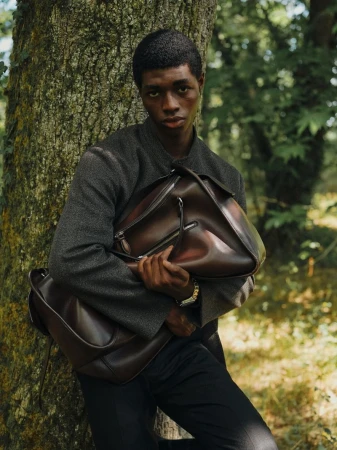 Man in grey jacket holds brown leather bag in forest. Portrait of adult male with brown hair and dark clothing in natural setting.