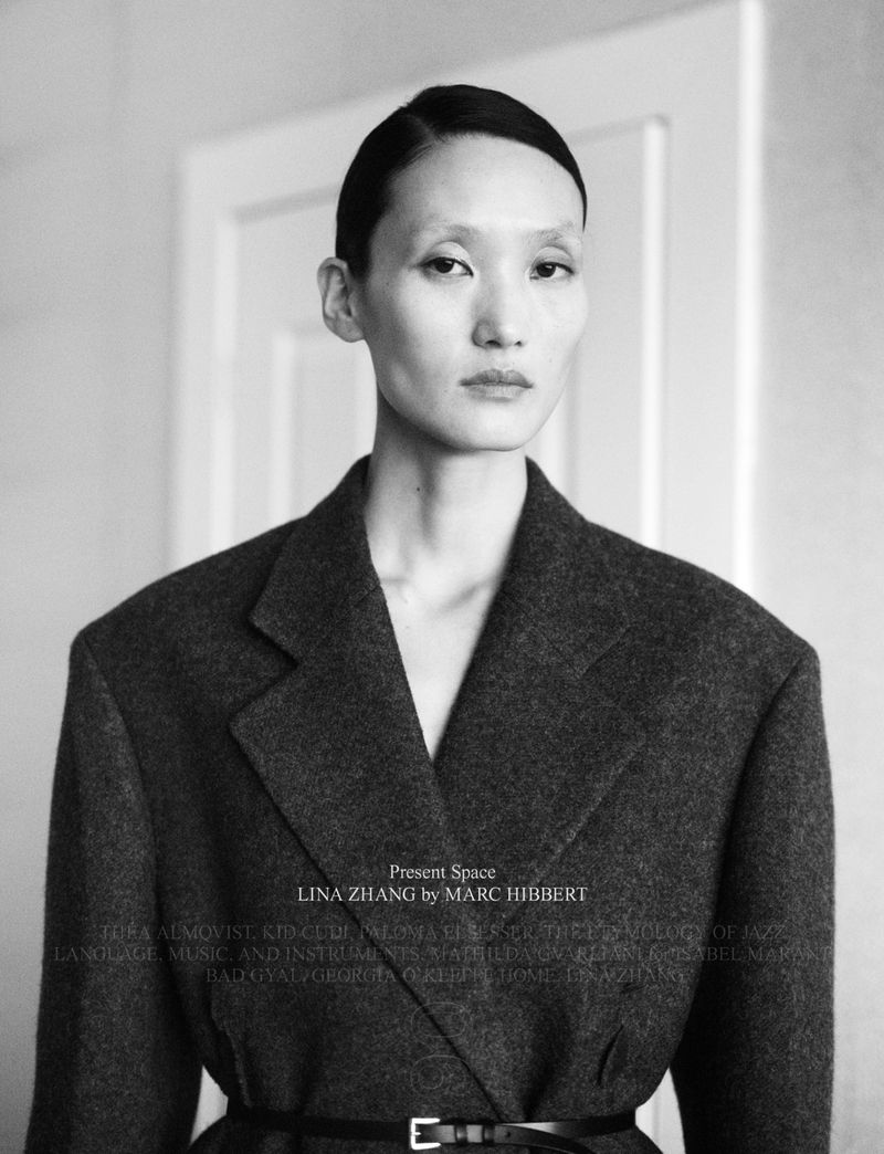 Portrait of an Asian woman with short dark hair wearing a grey coat with a black belt, standing in an indoor setting in a black and white image.