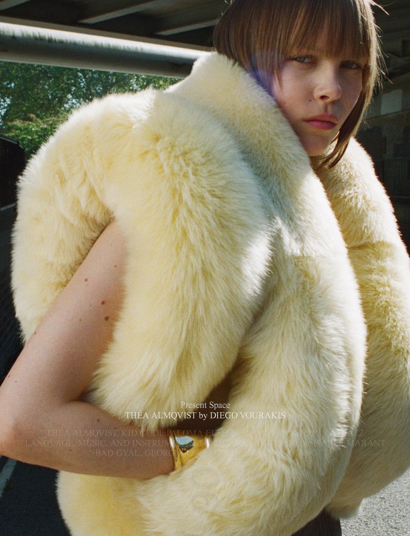 A woman in a cream-colored, furry shawl poses in an urban setting with a gold cuff bracelet and short, light-brown hair.