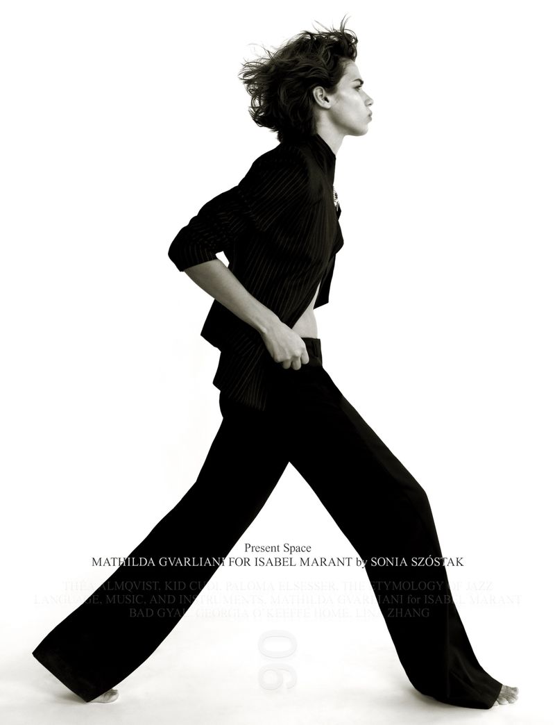 A person poses in dark pinstripe suit with flared pants in a studio shot against white backdrop, showcasing fashion and style.